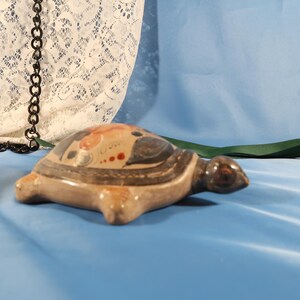 Może przedstawiać: Ceramiczna figurka żółwia z malowanym pancerzem z czerwonymi, niebieskimi i beżowymi wzorami. Żółw ma brązowe ciało i głowę i spoczywa na niebieskiej powierzchni. Figurka jest przedmiotem dekoracyjnym.