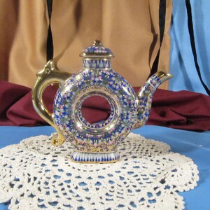 Puede incluir: Una tetera de cerámica decorativa con un diseño circular único en el centro. La tetera presenta un fondo azul con patrones florales en rosa, blanco y verde. Tiene un asa y un pico dorados, y está sobre un tapete de encaje blanco.