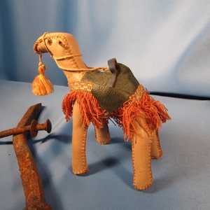 May include: A small, tan leather camel figurine with a green leather saddle and orange fringe. The camel has a blue leather strap around its neck and a long orange tassel.