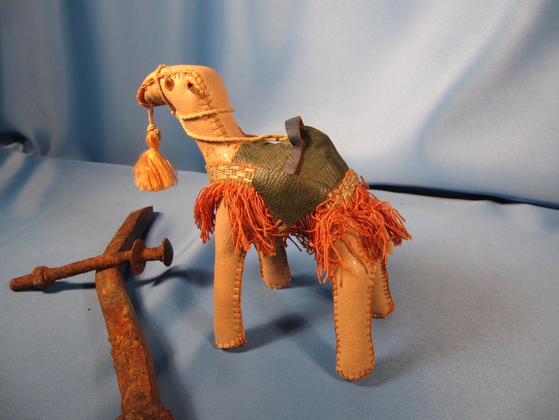 May include: A small, tan leather camel figurine with a green leather saddle and orange fringe. The camel has a blue leather strap around its neck and a long orange tassel.