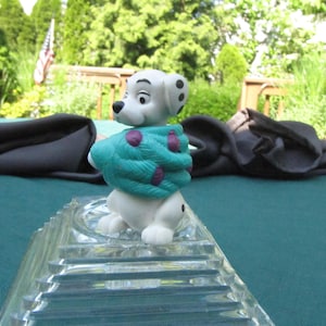 Może przedstawiać: Figurkę dalmatyńczyka w biało-czarnym kolorze, ubraną w turkusowo-fioletowy szalik. Figurka stoi na przezroczystym szklanym cokole.