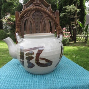 May include: A white ceramic teapot with a brown handle and brown and blue painted designs. The teapot is sitting on a blue and white patterned surface.