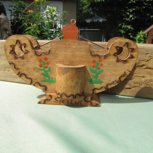 May include: Wooden candle holder with a carved design and painted flowers. The holder has a small, round top and a base with the word "Sicily" written on it.