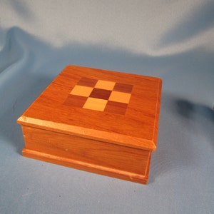 May include: A small wooden box with a checkered inlay on the top. The box is made of light and dark wood and has a natural finish.