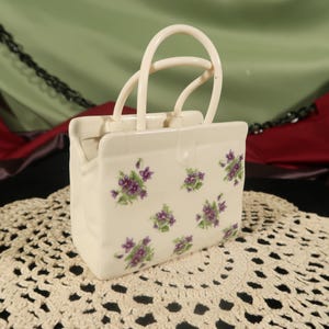 Puede incluir: Un bolso de mano de cerámica blanca con un diseño floral con flores moradas. El bolso tiene un asa y es un artículo decorativo.