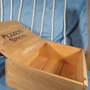 May include: A light brown wooden box with the words "PENZEYS SPICES" stencilled on the lid. The lid is slightly ajar, revealing the empty interior. The box is made of light-coloured wood and has a simple, rectangular design.