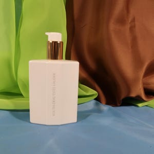 May include: A white rectangular bottle with a gold pump dispenser. The bottle has the text "YOUTH-DEW ESTEE LAUDER" printed vertically on the side. The background features green, brown, and blue fabric.
