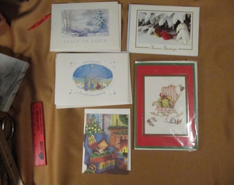 May include: Five Christmas cards with winter scenes. The cards feature a snowy landscape, a cardinal, a nativity scene, a fireplace, and a rocking chair with a gift. The cards have the text "Peace on Earth", "Season's Greetings", "Joy to the World", and "God could never love you more".