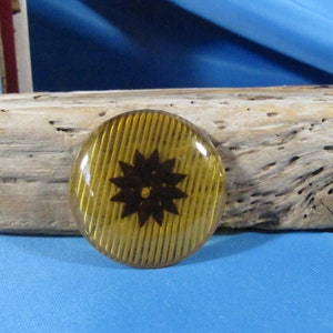May include: A round, yellow button with a black flower design. The button has a striped pattern.