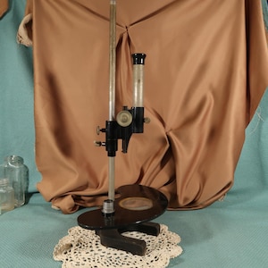 May include: A vintage black and silver microscope with a glass tube and a round black base. The microscope is on a white doily and a blue surface.