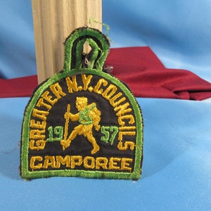 May include: A green, black, and gold embroidered patch with the text "GREATER N.Y. COUNCILS 1957 CAMPOREE". A cartoon figure of a person with a backpack and a walking stick is in the center of the patch.
