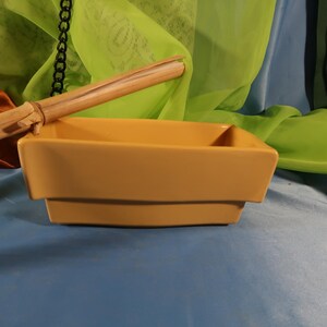 May include: A square, mustard-yellow ceramic bowl with a tiered design. A light brown wooden stick rests on the bowl's edge. The background features draped fabric in shades of green, blue, and brown. The bowl is likely for decorative or functional use.