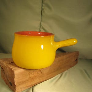 May include: A small yellow ceramic pot with a red rim and a handle. The pot is sitting on a piece of wood.