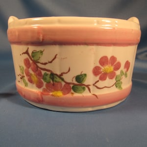 May include: A white ceramic bowl with pink and green floral designs. The bowl has a scalloped rim and two handles.