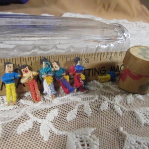 Puede incluir: Un conjunto de seis pequeñas muñecas de lana hechas a mano con cabezas y extremidades de madera. Las muñecas están dispuestas en fila sobre un mantel de encaje blanco. También se puede ver una regla de madera y una pequeña caja redonda decorada en la imagen.
