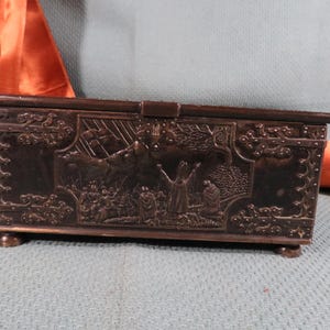 Puede incluir: Caja decorativa rectangular de color marrón oscuro con tapa abisagrada. La caja presenta tallas en relieve detalladas que representan una escena religiosa con figuras y un paisaje. La caja descansa sobre cuatro pequeños pies redondeados.