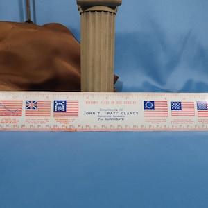 Puede incluir: Una regla vintage con banderas americanas históricas. La regla blanca tiene banderas impresas a lo largo de su longitud, con medidas en centímetros. El texto en la regla dice "Historic Flags of Our Country" y "John T. 'Pat' Clancy For Surrogate."