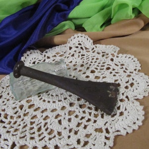 May include: A black metal tool with a long handle and a flat, rectangular head, resting on a white crocheted doily.