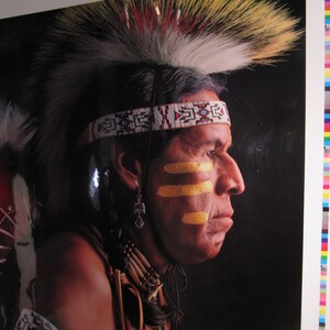 May include: A Native American man wearing a traditional headdress with feathers and a beaded headband. He has yellow face paint and a serious expression.