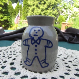 May include: A small gray ceramic jar with a blue gingerbread man design. The gingerbread man is wearing a blue bow tie and has blue buttons.