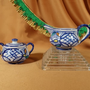 May include: A blue and white ceramic sugar bowl with a lid and two handles, and a matching blue and white ceramic creamer with a handle. Both pieces have a geometric pattern with yellow and green accents.