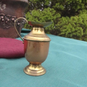 May include: A small, brass pitcher with a rounded body and a handle. The pitcher has a shiny, polished finish and a slightly aged patina.