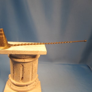 May include: A gold metal bar spoon with a decorative, engraved, cone-shaped end. The spoon is twisted and has a long, thin handle.