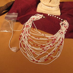 May include: A long, multi-strand necklace made of white and red beads. The necklace is laid out on a red fabric surface.