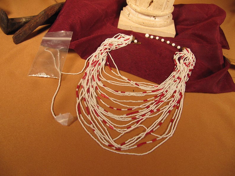 May include: A long, multi-strand necklace made of white and red beads. The necklace is laid out on a red fabric surface.