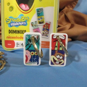 May include: A yellow box of SpongeBob SquarePants Dominoes with the Nickelodeon logo. Two white dominoes with gold key rings are in the foreground, featuring SpongeBob and Squidward. A stack of gold key rings is in the lower left.