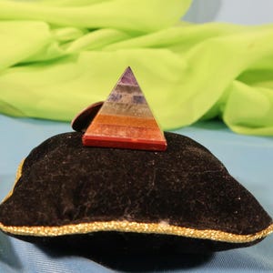 May include: A layered gemstone pyramid, featuring colors from red to purple, rests on a black velvet cushion with gold trim. The pyramid is a decorative item, possibly for meditation or display, against a backdrop of green fabric and a blue surface.