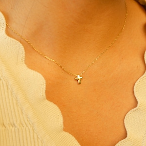 May include: A delicate gold-colored necklace featuring a small cross pendant. The cross is centered on a fine chain, creating a minimalist and elegant design. The necklace is worn against a light-colored top.