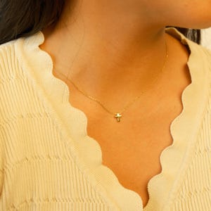 May include: A delicate gold necklace featuring a small cross pendant. The necklace is displayed against a light yellow top, highlighting the pendant's subtle design. The chain is fine, adding to the necklace's understated elegance.