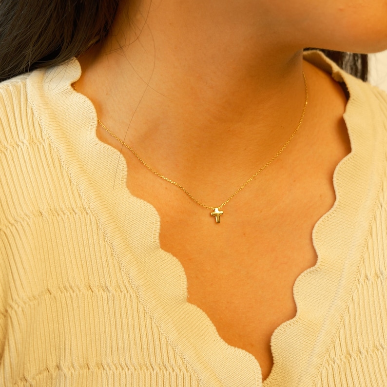 May include: A delicate gold necklace featuring a small cross pendant. The necklace is displayed against a light yellow top, highlighting the pendant's subtle design. The chain is fine, adding to the necklace's understated elegance.