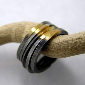 May include: A stack of five rings, four are silver and one is gold. The rings are stacked on a piece of driftwood.