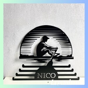 Puede incluir: Colgador de medallas de metal negro con una silueta de una persona remando un bote frente a una puesta de sol. El colgador tiene una placa de nombre que dice "NICO".