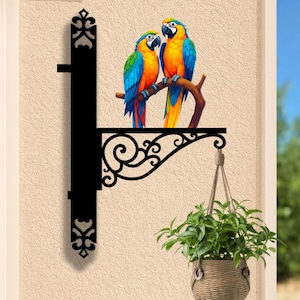 May include: Black metal hanging bracket with a decorative scroll design, featuring a colorful illustration of two macaws perched on a branch. A woven basket planter with green foliage hangs below. The text "LEGENDNADE" is at the bottom.