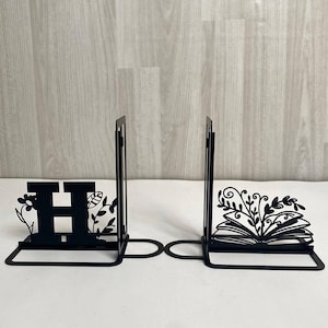Puede incluir: Soportes para libros de metal negro. Un sujetalibros presenta la letra "H" con detalles florales, y el otro representa un libro abierto con hojas decorativas. La palabra "LEGENDNADE" está impresa en la parte superior.