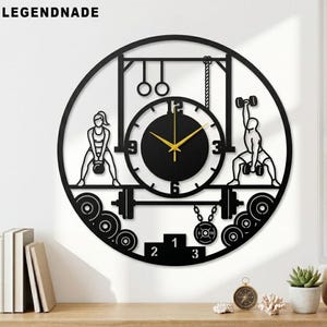 Może przedstawiać: Czarny metalowy zegar ścienny o tematyce fitness. Zegar przedstawia sylwetki ciężarowców, sztangi i kettlebell. Tarcza zegara jest czarna ze złotymi wskazówkami. U góry znajduje się napis "LEGENDNADE".