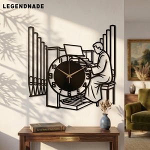 Op de afbeelding: Een zwarte metalen wandklok met een silhouet van een persoon die op een orgel speelt. De wijzerplaat is rond met gouden wijzers en zwarte cijfers. Het ontwerp bevat verticale orgelpijpen en de tekst LEGENDNADE in de bovenhoek.