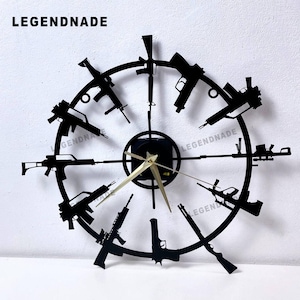 Puede incluir: Reloj de pared de metal negro con siluetas de armas de fuego en la esfera. El reloj tiene manecillas doradas y el texto "LEGENDNADE" en la parte superior. El diseño incorpora varias formas de armas de fuego.