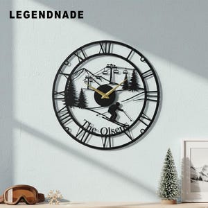 May include: Black metal wall clock with a ski resort scene. The clock features Roman numerals, gold-colored hands, and the text "The Olsens". The clock is round and has a ski lift, trees, and a skier silhouette.