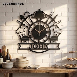 Könnte beinhalten: Eine Wanduhr aus schwarzem Metall mit einem zentralen runden Zifferblatt, römischen Ziffern und goldfarbenen Zeigern. Das Design zeigt Küchenmotive wie Kochmützen, Servierplatten und Besteck. Der Name JOHN ist auf einem Banner zu sehen.