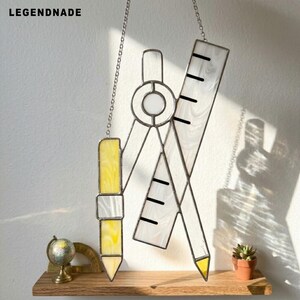 May include: A stained glass suncatcher featuring a yellow pencil, a drafting compass, and a ruler, all connected by a metal chain. The piece includes the text LEGENDNADE at the top left. The glass has a marbled white and yellow texture.