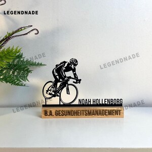 May include: A black and white cyclist silhouette on a bicycle, mounted on a wooden base. The base has the name "NOAH HOLLENBORG" and "B.A. GESUNDHEITSMANAGEMENT" engraved on it. The product is a decorative item.