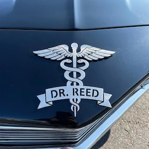 May include: A silver caduceus emblem with the text "DR. REED" on a black car hood. The emblem features two snakes entwined around a staff with wings at the top. The car's chrome grill is visible below the emblem.