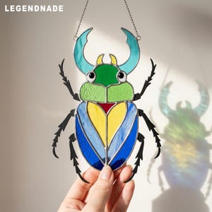May include: A stained glass suncatcher shaped like a beetle with the text LEGENDNADE. The insect features a green head, light green thorax, and wings with yellow and blue segments. It has black metal legs and antennae, suspended by a silver chain.