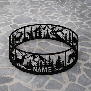 Puede incluir: Anillo de fogata de metal negro con una escena de bosque recortada con ciervos, árboles y montañas. La palabra "NAME" está cortada en la parte inferior del anillo. El anillo es circular y se encuentra sobre una superficie de piedra.