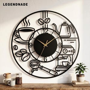 May include: A circular black metal wall clock featuring the text LEGENDNADE at the top. The design includes cut-out silhouettes of a coffee cup, coffee beans, a kettle, an espresso machine, and a portafilter. It has a central black dial with gold hands.