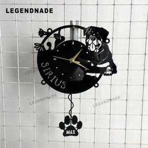 Puede incluir: Reloj de pared de metal negro con silueta de perro, la palabra "SIRIUS" y manecillas doradas. El reloj tiene un péndulo en forma de pata con el nombre "MAX". La marca "LEGENDNADE" es visible.